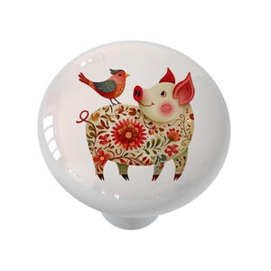May include: White ceramic cabinet knob featuring a whimsical illustration of a pig adorned with floral patterns and a perched bird. The pig has pink ears and a smiling face. The knob is a decorative accent for furniture.