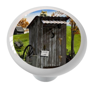 May include: A white cabinet knob featuring a rustic outhouse scene. The image includes a raccoon, a sign that reads "1 Hour Limit", and a crescent moon cutout. The background shows a green field and a small building.