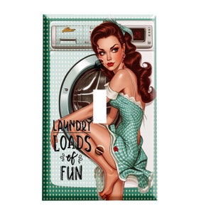 May include: A light switch cover featuring a vintage illustration of a woman in a green dress leaning against a white washing machine with the text "Laundry Loads of Fun". The background is a green and white polka dot pattern.