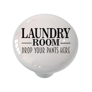 May include: A white ceramic cabinet knob with the text "LAUNDRY ROOM - DROP YOUR PANTS HERE" in black. The knob has a glossy finish and a rounded shape, suitable for a laundry room.