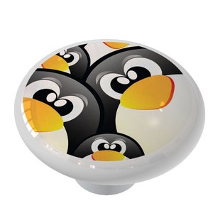 May include: A white ceramic knob with a colorful cartoon design of penguins with black and white bodies, orange beaks, and black eyes.
