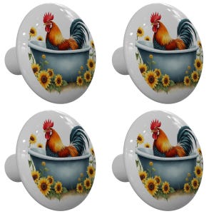 May include: Four white cabinet knobs with a colorful rooster design. Each knob features a rooster in a blue tub surrounded by sunflowers. The rooster has red, orange, and blue feathers. A decorative accent for furniture.