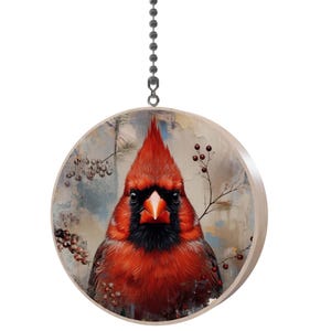 May include: A round wooden ornament featuring a detailed painting of a red cardinal bird. The bird has a black face and a prominent crest. The background has a soft, muted color palette with berry branches and a hanging chain.