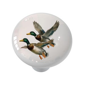 May include: White ceramic cabinet knob featuring a detailed illustration of two mallard ducks in flight. The ducks have green heads, brown bodies, and blue and gray wings. The knob has a smooth, glossy finish.