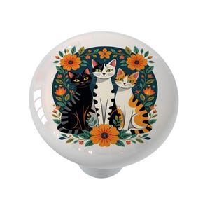 May include: White ceramic cabinet knob featuring a colorful illustration of three cats surrounded by orange flowers and green leaves. The cats are black, white, and orange, set against a dark blue background.