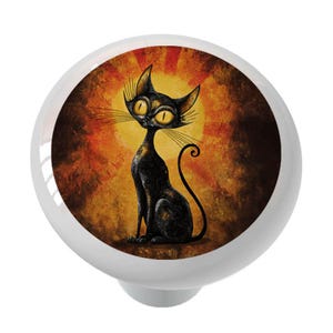 May include: A white cabinet knob featuring a black cat illustration with yellow eyes and a sunburst background. The cat is sitting upright with a curled tail. The knob is round with a small, white base.