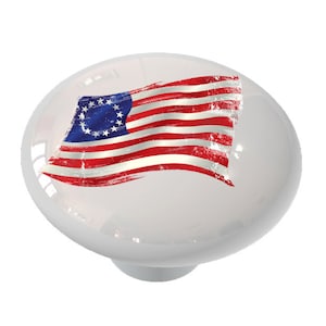 May include: A white ceramic knob with a distressed American flag design featuring 13 stars in a circle. The flag is red, white, and blue.