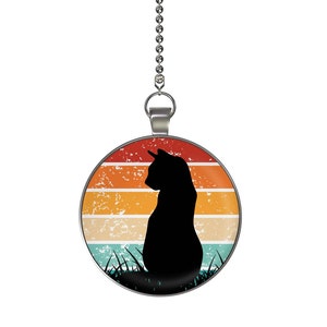 May include: A silver chain with a round pendant featuring a black silhouette of a cat sitting in front of a retro sunset with orange, yellow, and blue stripes.