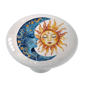 May include: A white ceramic knob with a watercolor illustration of a crescent moon and sun. The moon is blue with stars and the sun is yellow with a face.