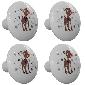 May include: Four white ceramic cabinet knobs with a festive design. Each knob features a brown reindeer with a red nose, a black and white scarf, and snowflakes. The knobs are perfect for adding a touch of holiday cheer to any cabinet or drawer.