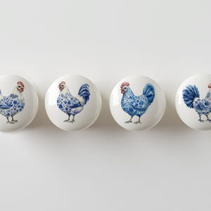 May include: Four white ceramic cabinet knobs with a blue and white rooster design. Each knob features a different rooster illustration with a red comb and blue floral patterned feathers. The knobs are round and have a glossy finish.