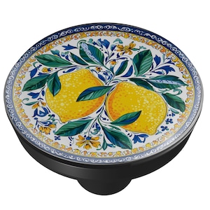 May include: A round, black bottle stopper with a decorative top featuring a lemon design. The top has a white background with yellow lemons, green leaves, and blue floral accents. The rim has a blue and yellow pattern.