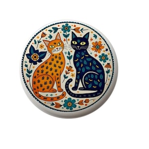 May include: A white ceramic plate with a colorful floral design featuring two cats. One cat is orange with black spots and the other is blue with green spots. The cats are sitting side by side and looking at each other.