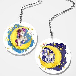 May include: Two white round fan pull charms with a unicorn on a yellow crescent moon against a blue and black starry night background.