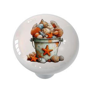 May include: White ceramic cabinet knob featuring a watercolor illustration of a bucket overflowing with seashells and starfish. The bucket is light green with orange accents. The seashells are orange, white, and blue.