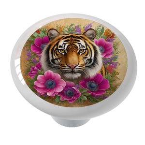 May include: White ceramic cabinet knob featuring a tiger's face surrounded by pink flowers and greenery. The tiger has orange and black stripes with a tan background. The knob is a decorative accent for furniture.