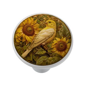 May include: A round cabinet knob featuring a detailed illustration of a yellow bird perched on a branch surrounded by two large, golden-yellow sunflowers with dark brown centers and green leaves, set against a circular white base.