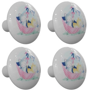 May include: Four white ceramic cabinet knobs. Each knob features a watercolor illustration of two kittens under a pink polka-dot umbrella with a blue sky background and a small bird. The knobs have a rounded shape.