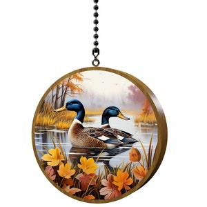 May include: A round wooden ornament with a picture of two ducks swimming in a lake with fall leaves. The ducks are brown, blue, and white. The lake is blue and the leaves are orange, yellow, and red.