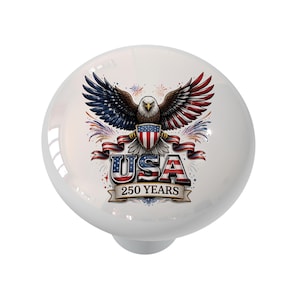 May include: White ceramic cabinet knob featuring a patriotic design. The design includes a detailed illustration of an American bald eagle with outstretched wings, the word "USA", and the text "250 YEARS" on a banner.