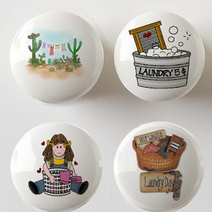 May include: Four white ceramic cabinet knobs featuring distinct illustrations: a desert scene with a clothesline, a metal washbasin with bubbles and the text LAUNDRY 5¢, a cartoon character with a laundry basket, and a basket labeled Laundry Day.