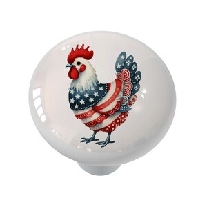 May include: A white ceramic cabinet knob featuring a colorful rooster design. The rooster is decorated with red, white, and blue patterns, including stars and stripes, reminiscent of the American flag. The knob has a glossy finish and a round shape.