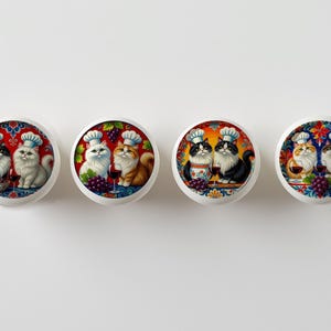 May include: Four white ceramic cabinet knobs with colorful illustrations of cats dressed as chefs. Each knob features a pair of cats, grapes, and wine glasses. The cats are in various colors, including black, white, and orange.