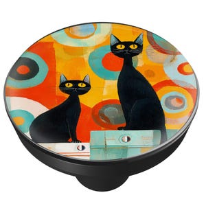 May include: A black PopSocket with a colorful abstract design featuring two black cats with yellow eyes. The background has orange, yellow, and blue circles. The cats are sitting on a light blue surface.