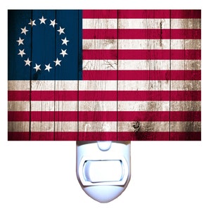 May include: A white night light with a red, white, and blue American flag design. The flag has 13 stripes and a circle of 13 stars on a blue background. The flag is on a woodgrain background.