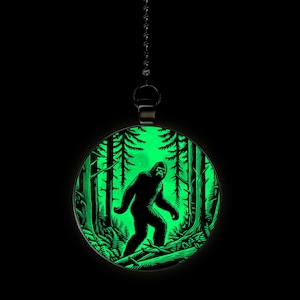 May include: A glow-in-the-dark pendant featuring a silhouette of Bigfoot walking through a forest. The pendant is round and has a black metal chain.
