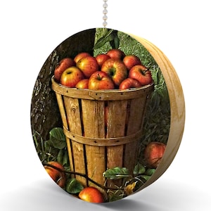 May include: A wooden round pull chain with a picture of a basket full of red and yellow apples. The basket is made of wood and is sitting on a bed of green grass.