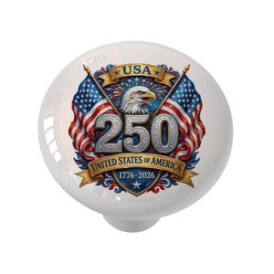 Puede incluir: Un pomo de armario redondo blanco con un diseño patriótico. El diseño incluye el texto "USA" y "250 UNITED STATES OF AMERICA 1776-2026" con un águila, banderas americanas y colores rojo, blanco y azul.