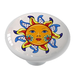 May include: A ceramic knob with a colorful sun design. The sun has a face with blue eyes, a red mouth, and yellow flowers on its cheeks. The sun is surrounded by blue, red, and yellow rays.