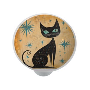May include: A decorative cabinet knob featuring a black cat illustration with teal eyes and star details. The cat is set against a tan background with teal starbursts. The knob has a white base.