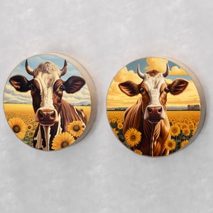 May include: Two round wall art pieces featuring cow portraits in a sunflower field. The cows have brown and white markings, with yellow sunflowers in the foreground and a blue sky background. The art pieces have a wooden frame.