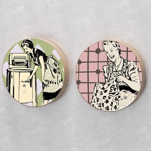 May include: Two round wooden coasters with vintage illustrations of women doing housework. One coaster features a woman in a dress and apron operating a vintage washing machine. The other coaster features a woman in a dress and apron sewing. Both coasters have a checkered background.
