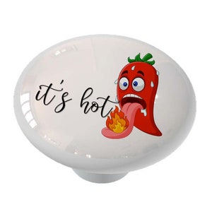 May include: White ceramic knob with a red chili pepper cartoon with a tongue sticking out and flames coming from the tongue. The text "it's hot" is written in black cursive.