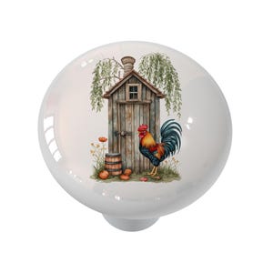 May include: A white ceramic cabinet knob featuring a watercolor illustration. The design includes a rustic wooden outhouse, a colorful rooster, a small barrel, and orange pumpkins. The knob has a glossy finish.