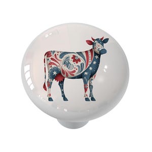 May include: A white ceramic cabinet knob featuring a decorative cow design. The cow is adorned with a patriotic pattern in red, white, and blue, including stars, flowers, and swirls. The knob has a glossy finish and a round shape.