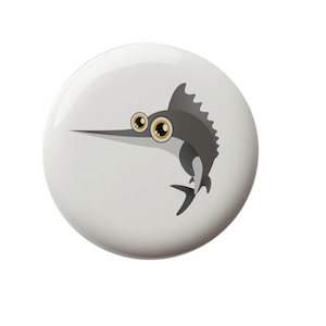 May include: A white button with a cartoon gray marlin with large yellow eyes. The marlin is swimming to the right.