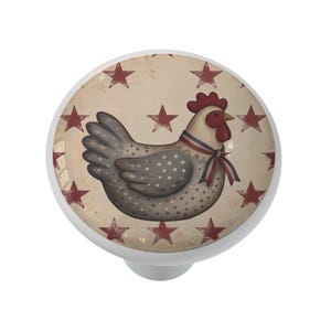 May include: A white cabinet knob with a decorative chicken design. The chicken is gray with white polka dots and a red comb, wearing a red, white, and blue ribbon. The background is beige with red stars.