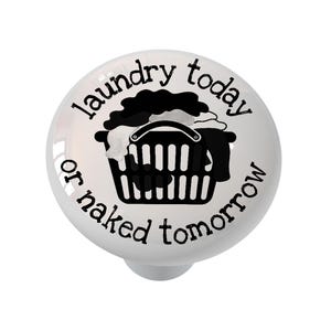 May include: A round white ceramic cabinet knob featuring a black graphic of a laundry basket filled with clothes. The text "laundry today or naked tomorrow" is printed in a black sans-serif font, curving around the central laundry basket illustration.