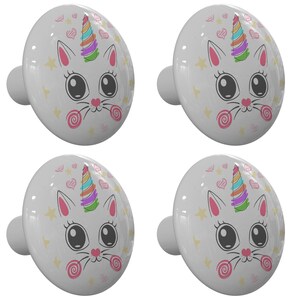 May include: Four white ceramic cabinet knobs with a whimsical design of a white cat with a rainbow unicorn horn, surrounded by pink hearts, yellow stars, and pink and white candy swirls.