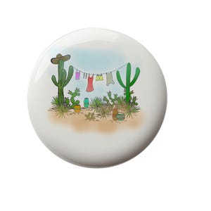 May include: A white ceramic plate with a cartoon illustration of a desert landscape with cacti and a clothesline with colorful clothing hanging on it.