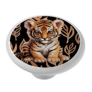 May include: A white cabinet knob featuring a detailed illustration of a baby tiger cub. The tiger has orange and black stripes, with a black background and gold leaf pattern. Decorative hardware for furniture.
