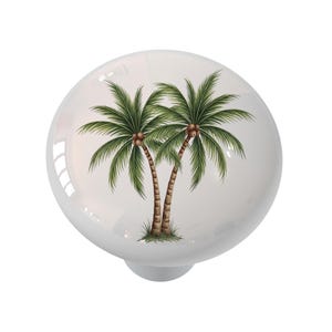 May include: A round white cabinet knob featuring a detailed illustration of two palm trees with green fronds and brown trunks. The design is centered on the smooth, glossy surface of the knob, which has a curved, dome-like shape and a solid white finish.