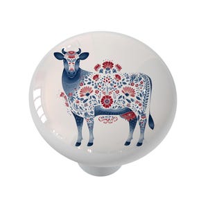 May include: A white ceramic cabinet knob featuring a detailed illustration of a cow. The cow is decorated with red and blue floral patterns. The knob has a glossy finish and a round shape.