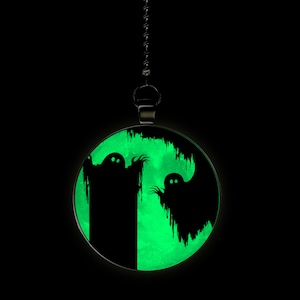 May include: A black and green glow-in-the-dark pendant featuring two ghosts. The pendant is hanging from a silver chain.
