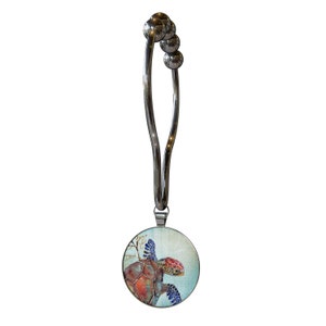 May include: A silver metal curtain tieback with a round, colorful sea turtle design. The turtle is swimming in blue water with coral and seaweed.