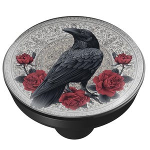 May include: A black PopSocket with a detailed illustration of a raven perched on a branch of red roses. The background features an ornate, off-white floral design. The base of the PopSocket is black.